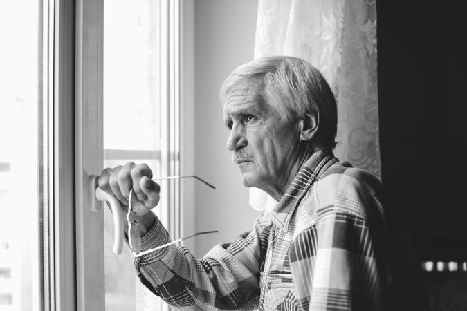 Side view of older man holding eyeglasses standing by the window in grayscale