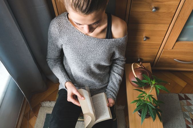 Woman relaxing with a book, pot of tea and marijuana plant