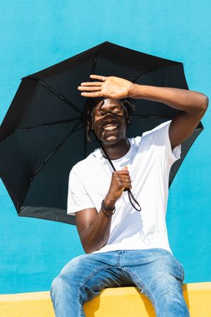Happy Black man with umbrella sitting on fence