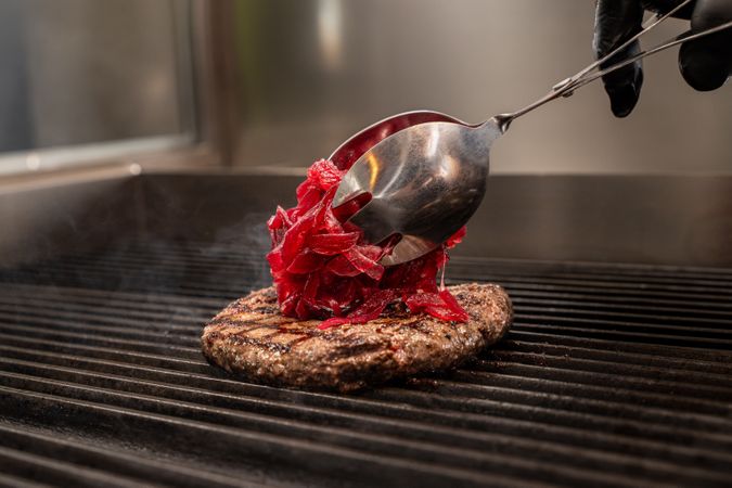 Chef adding caramelized red onions to grilled burger patty on barbecue