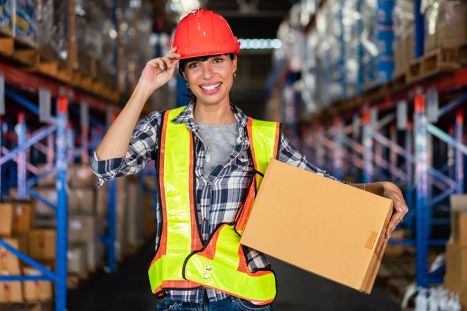 Smiling female warehouse worker with vision in safety gear holding a box