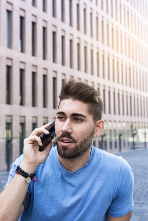 Portrait of man with beard in blue t-shirt using phone while sitting outside building