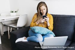 Long haired female sitting on sofa with laptop computer working remotely from home checking phone 48BlEk