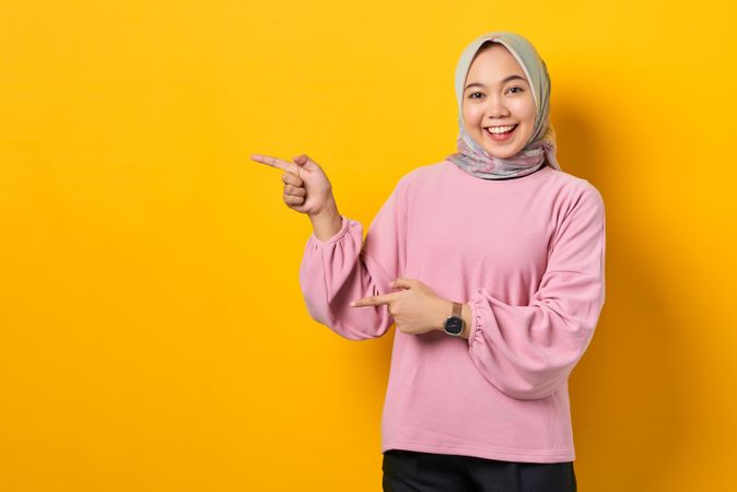 Happy Muslim woman in scarf and pink sweater pointing at copy space studio shoot