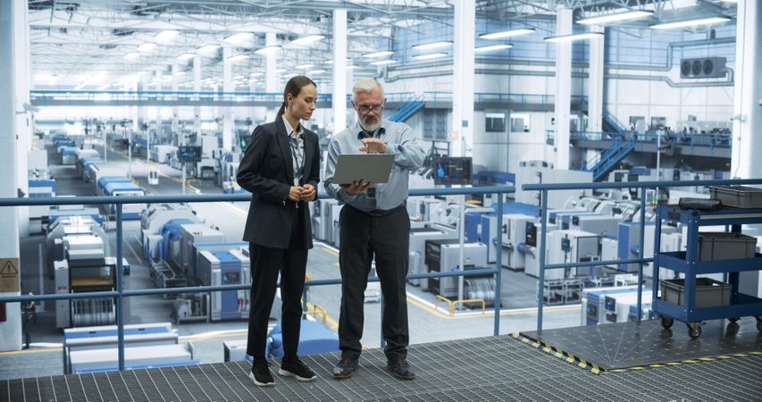 Mature Engineer and Young Female Technician Having a Conversation and Using Laptop Computer. Specialists Monitoring, Analyzing System Reports at a Modern Electronics Factory
