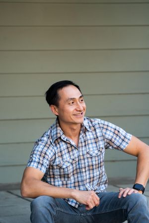 Confident man sitting in front of house wearing plaid shirt sitting and looking away