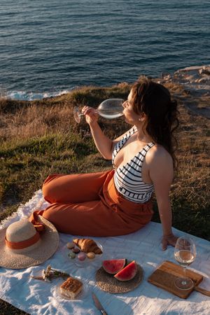 Woman with glass of drink on picnic mat looking at sea and mountains