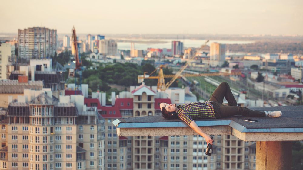 Male lying back looking at sky, with bottle of wine in hand on roof