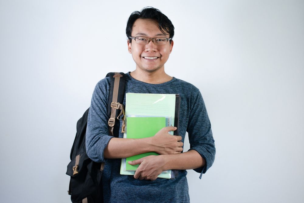 Asian young male student wearing bag and carrying books smiling and happy expression
