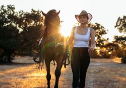 Young woman in hat walking with her horse in the countryside at sunset