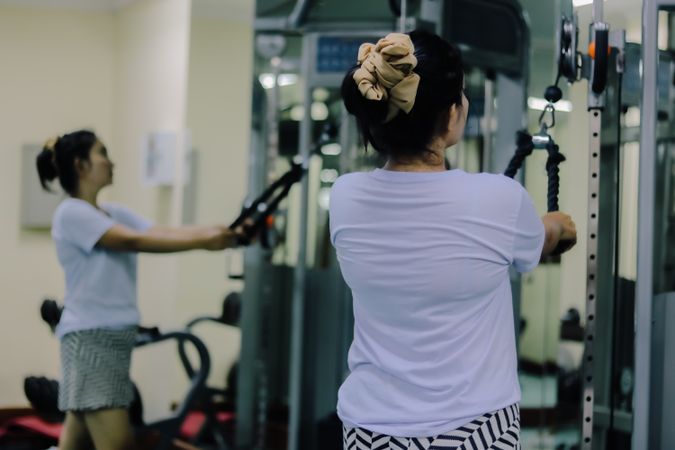 A woman with her back facing forward is using a cable machine, holding the handles, and exercising at a gym.