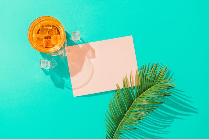 Tropical cocktail with ice cubes, palm leaf  and pink paper card note on vivid blue background