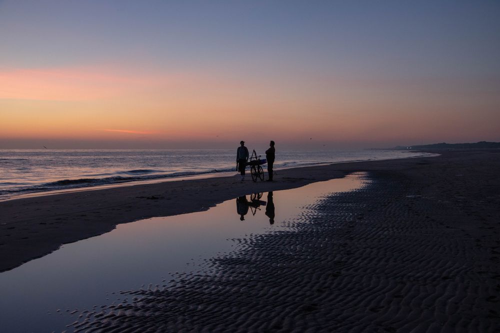 Silhouette of two people with bicycle standing on the beach at sunset
