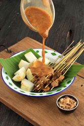 Peanut sauce being poured over plate of beef sate skewers