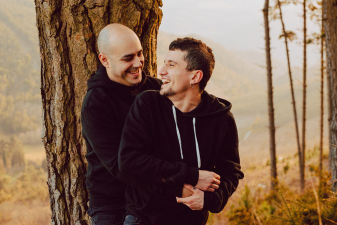 Smiling men hugging near tree in forest on hill
