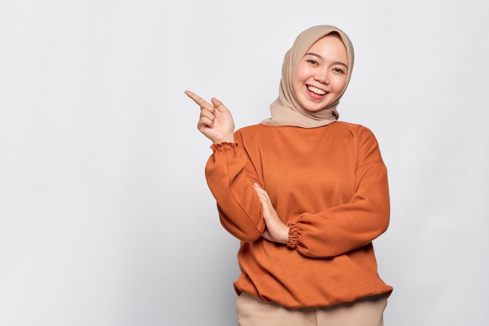 Smiling Muslim woman in headscarf and orange sweater pointing to her side