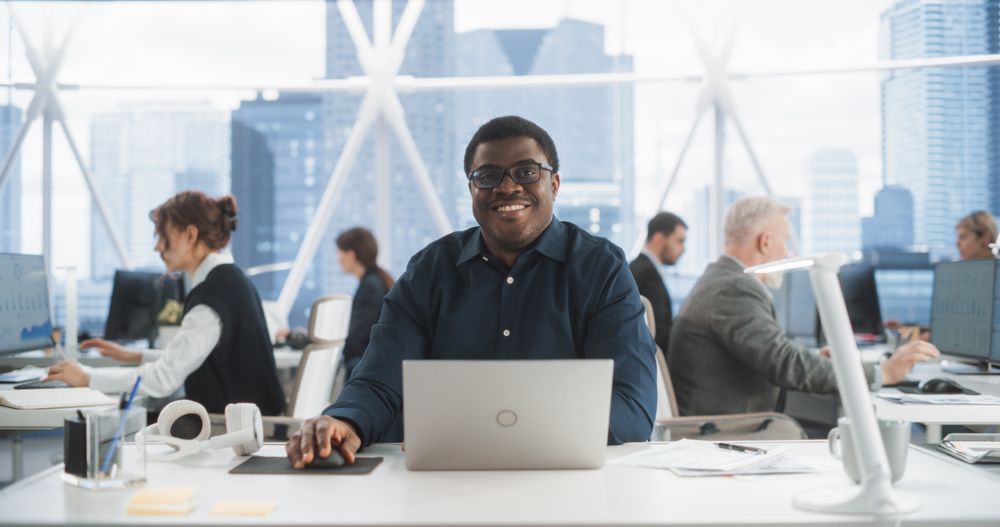 Successful Black Man Typing On Laptop Computer In Bright Skyscraper Office With Diverse Employees In Downtown.