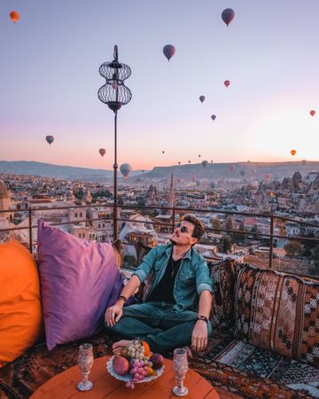 Man sitting on cushion on roof of building under sky filled with hot air balloon