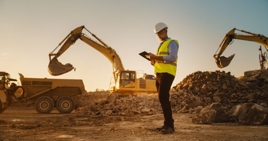Male Real Estate Investor Wearing Protective Goggles And Using Tablet On Construction Site On a Sunny Day. Man Inspecting Building Progress. Excavator Loading Materials Into Truck.
