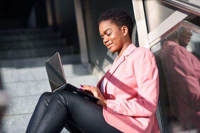 Female in pink suit working on shadowy stairs outside with laptop on knees