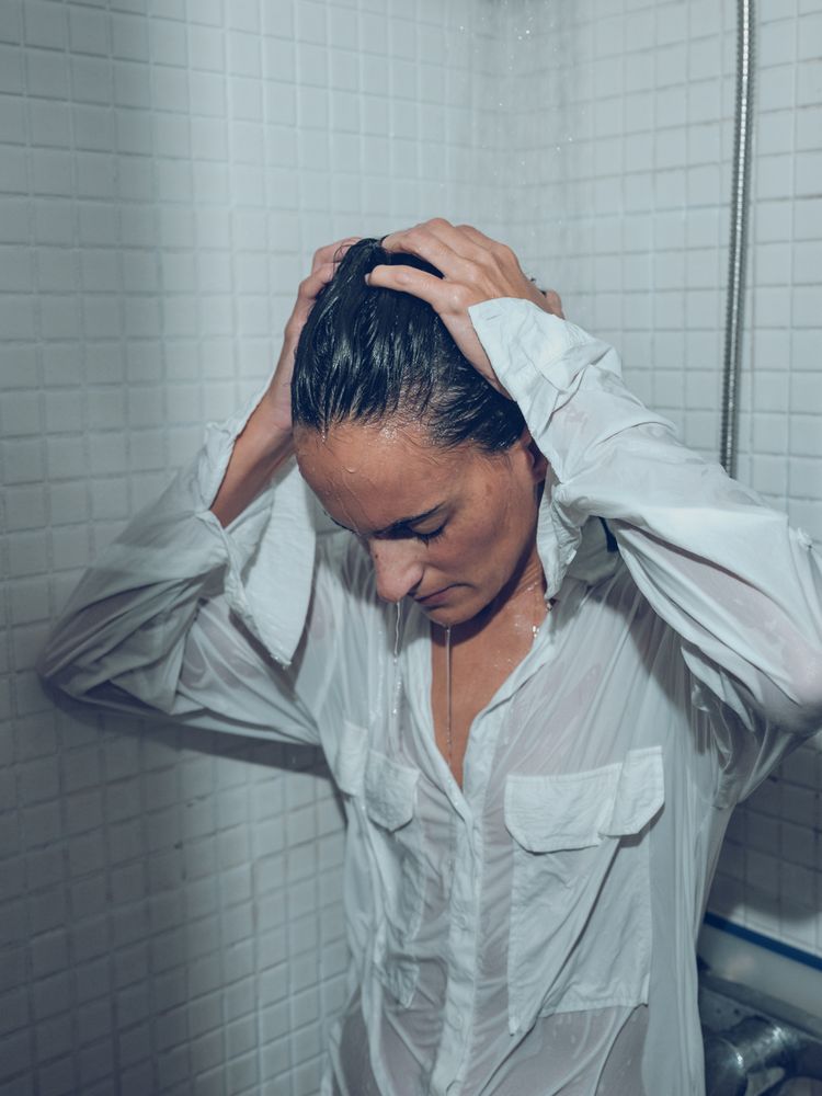 Exhausted woman in wet shirt touching head under shower