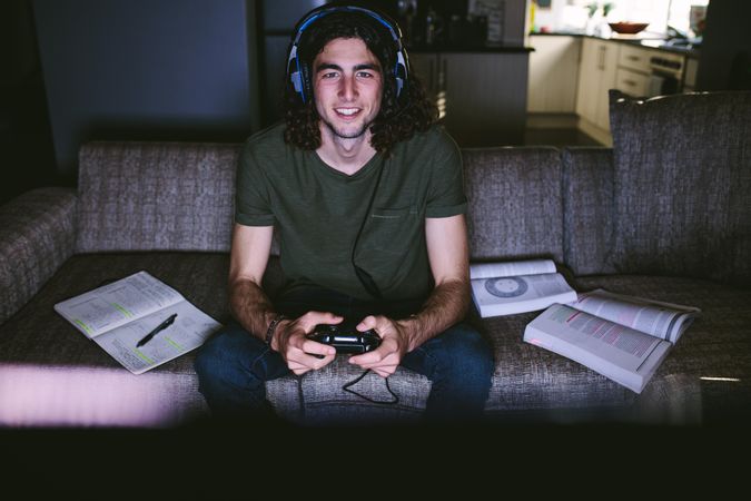 Student sitting on couch playing video game with books beside him