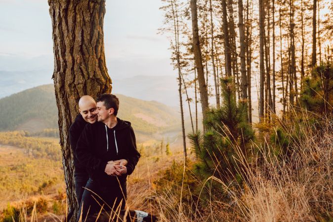 Smiling men hugging near tree in forest on hill