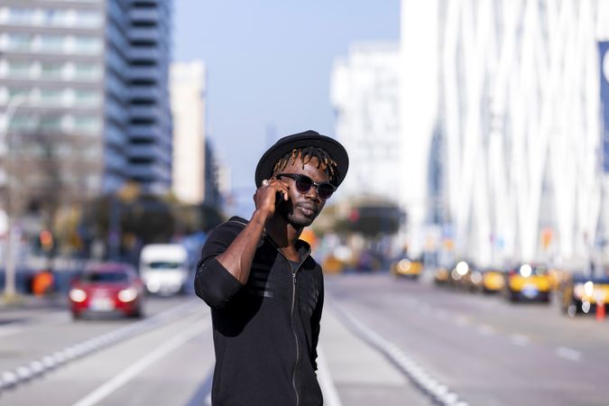 Black man wearing hat & sunglasses standing on the street using cellphone