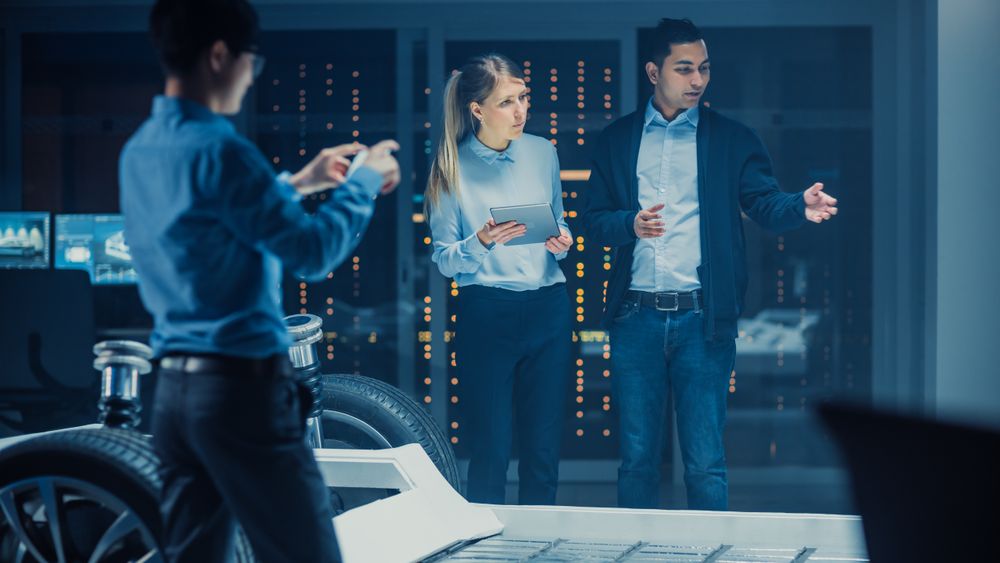 Shot of a Development Laboratory Room with Diverse Professional Team of Engineers and Scientists Working on a Electric Car Chassis with Wheels, Batteries, Engine and Suspension.