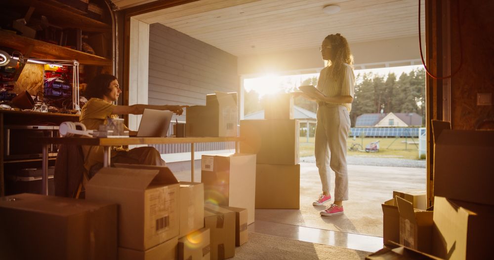 White Male Entrepreneur Using Laptop Computer To Fill Orders From Online Customers, Talking To Hispanic Female Business Partner With Tablet About Products Packaged In Cardboard Boxes For Delivery.