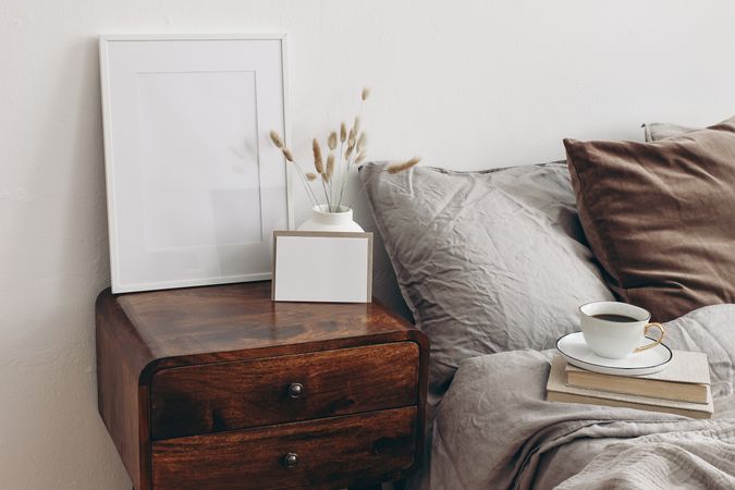 Elegant bedroom. Blank greeting card, invitation and picture frame mockup on old books. Brown, grey linen and velvet pillows. Wooden night stand with ceramic vase, dry grass. Cup of coffee. Scandinavian interior.
