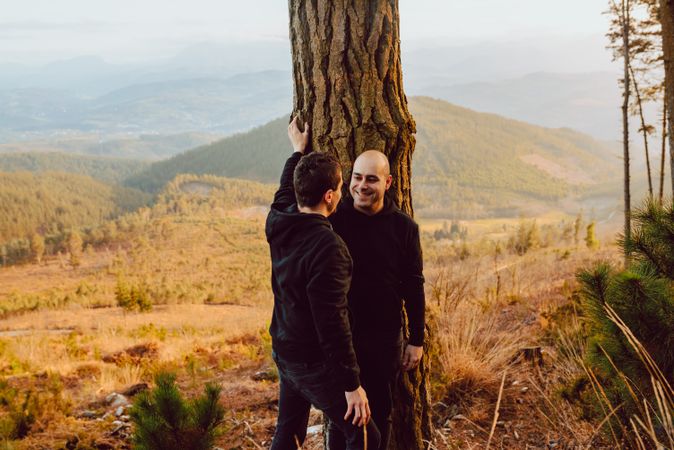 Smiling men hugging near tree in forest on hill