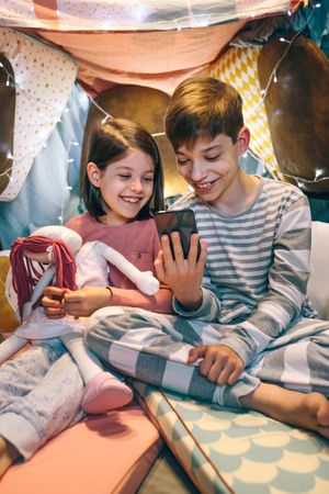 Happy children laughing together while watching video on cell phone sitting on shelter tent at home