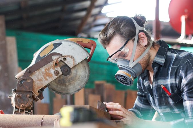 Male carpenter wearing protective mask using electric circular saw cutting wood board at workshop