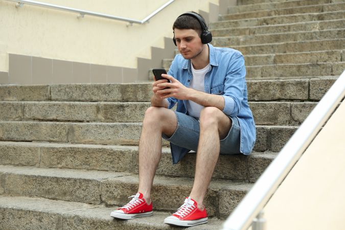 Young man in headphones sits on the steps and looking in phone