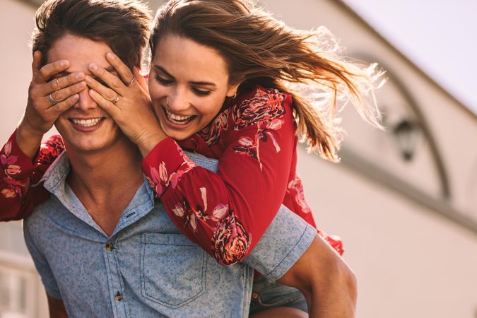 Smiling woman piggy back riding on man closing his eyes with her hands
