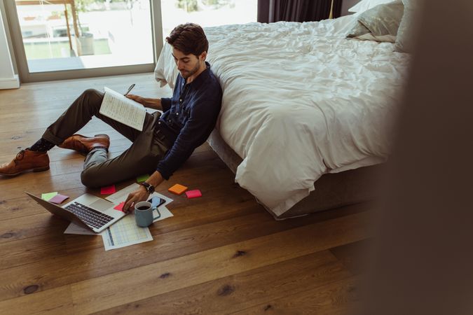 Man making notes in diary and looking at laptop sitting on bedroom floor