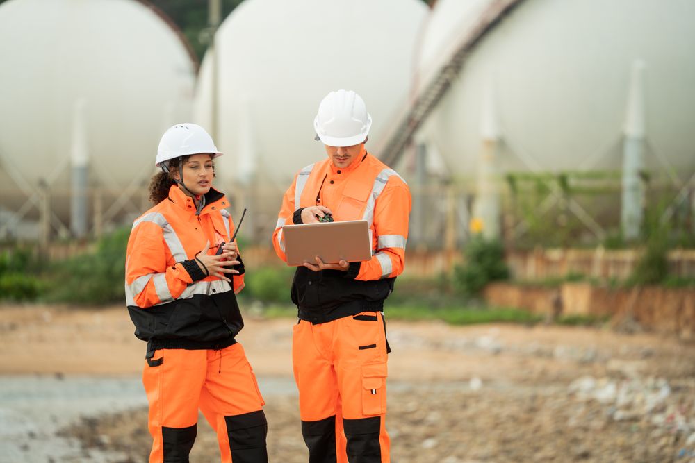 Two engineers in safety gear talking work near spherical gas storage tanks. woman is pointing forward while the man checks a laptop, indicating active discussion and site coordination in an industrial