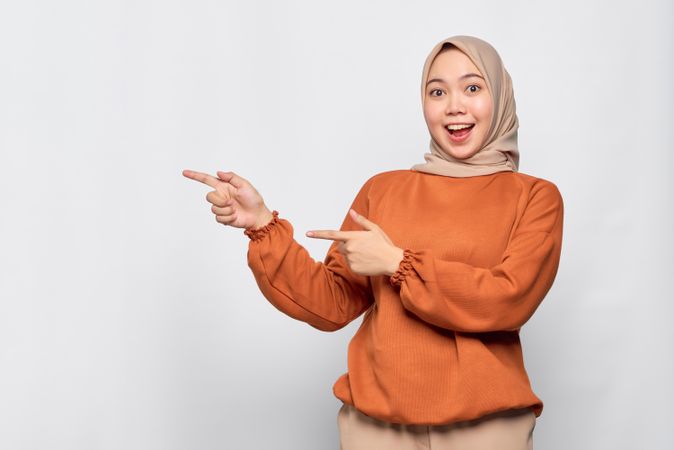 Enthusiastic Muslim woman in headscarf and orange sweater pointing to her side