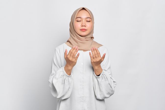 Muslim woman in headscarf and light blouse with eyes closed and hands up in prayer