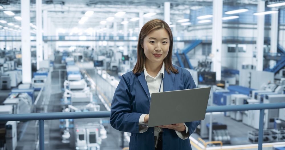 Portrait of an Electronics Manufacture Engineer Smiling, Looking at Camera. Young Japanese Female Using Laptop Computer for Online Research, Monitoring Artificial Intelligence Systems