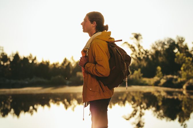 Side view of a man wearing backpack standing beside a lake and looking away