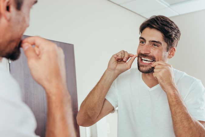 Man looking in the bathroom mirror and using dental floss to clean his teeth