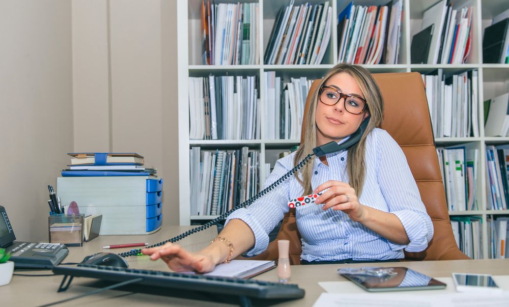 Blonde woman taking call, using computer and holding a nail file in office