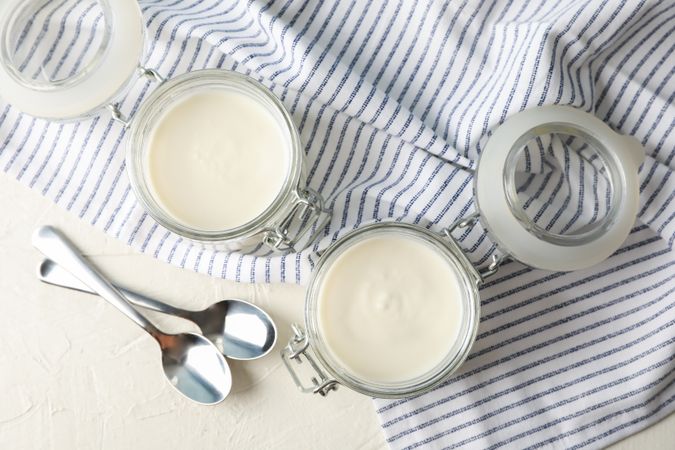 Glass jars with sour cream yogurt, spoons and towel on cement background