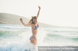 Side shot of woman in bikini jumping with outstretched arms and smiling in the water at the beach 5nQnA0