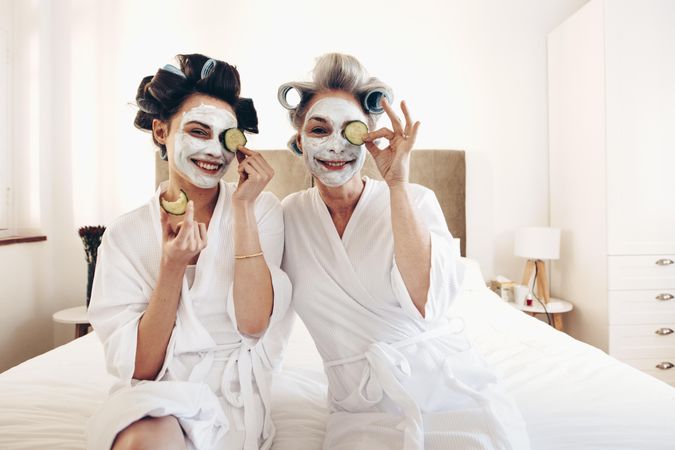 Smiling mother and daughter with beauty face mask sitting on bed holding slices of cucumber to eyes
