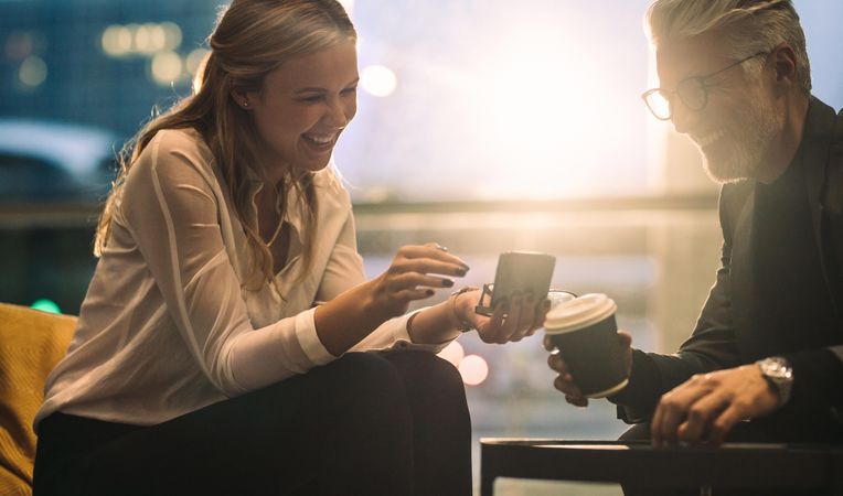 Smiling woman with colleague sitting in office lobby using mobile phone during coffee break