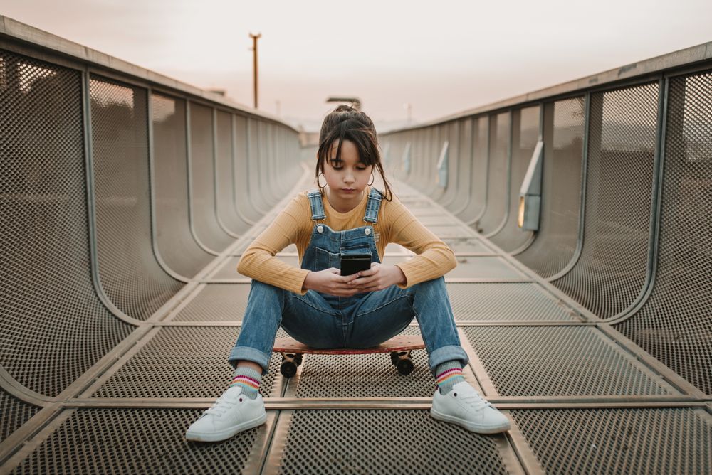 Girl with smartphone sitting on skateboard on walkway
