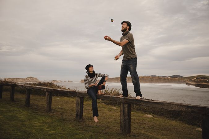 Man juggling balls on bench near woman with drum on shore